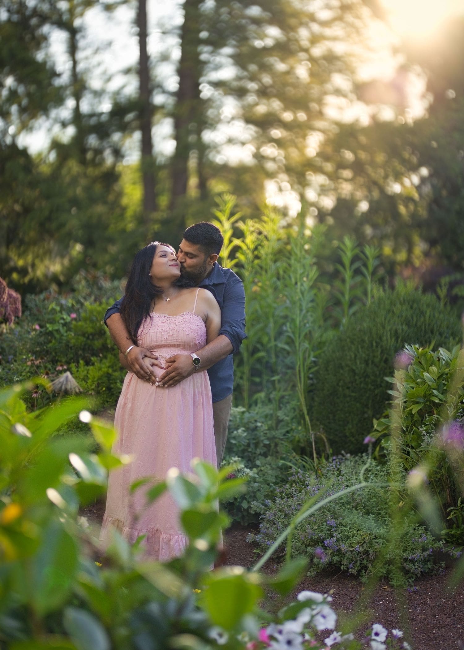 Expecting couple in soft pink and neutral outfits in a garden at golden hour